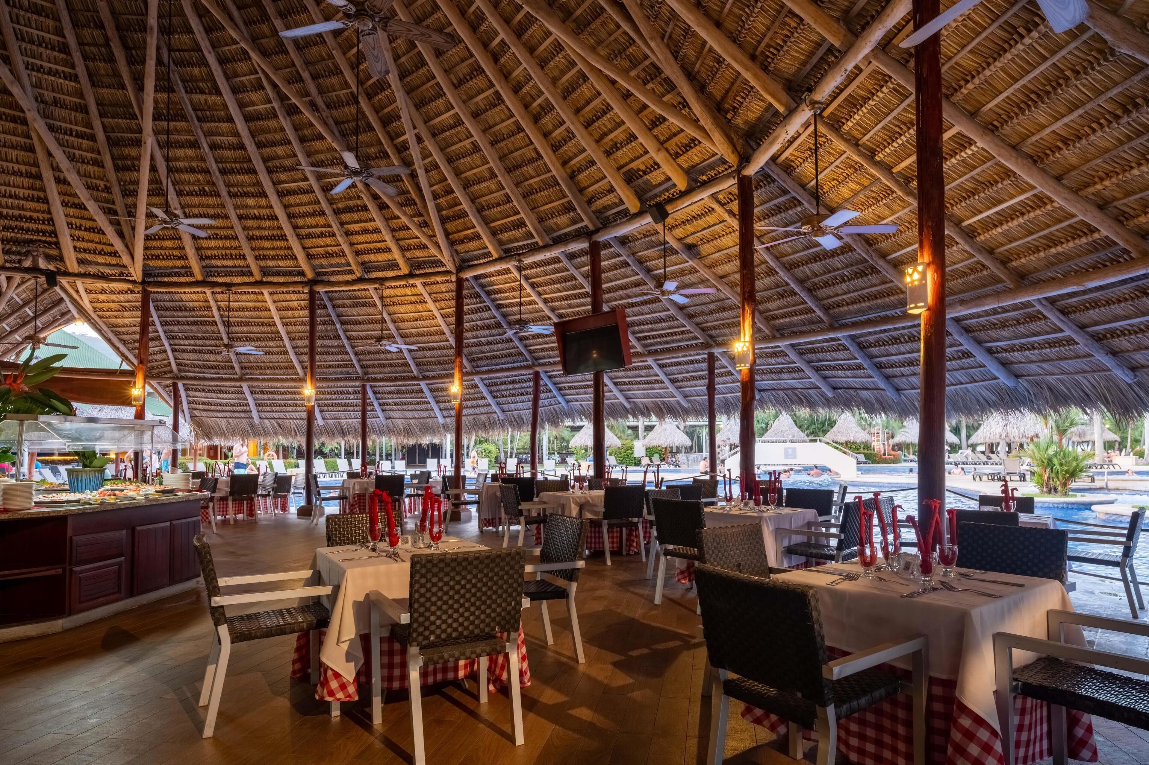 An outdoor restaurant with thatched roof, tables with red and white checkered tablecloths, and a buffet area.