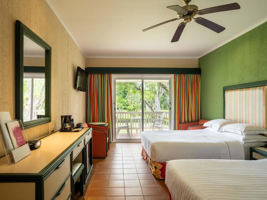 A hotel room with two beds, a desk with a mirror, a ceiling fan, and a balcony with colorful curtains.