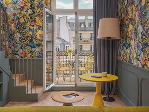 Un appartement moderne avec un papier peint floral aux motifs vifs, une table ronde jaune avec une lampe, et un balcon agrémenté de fauteuils en osier.