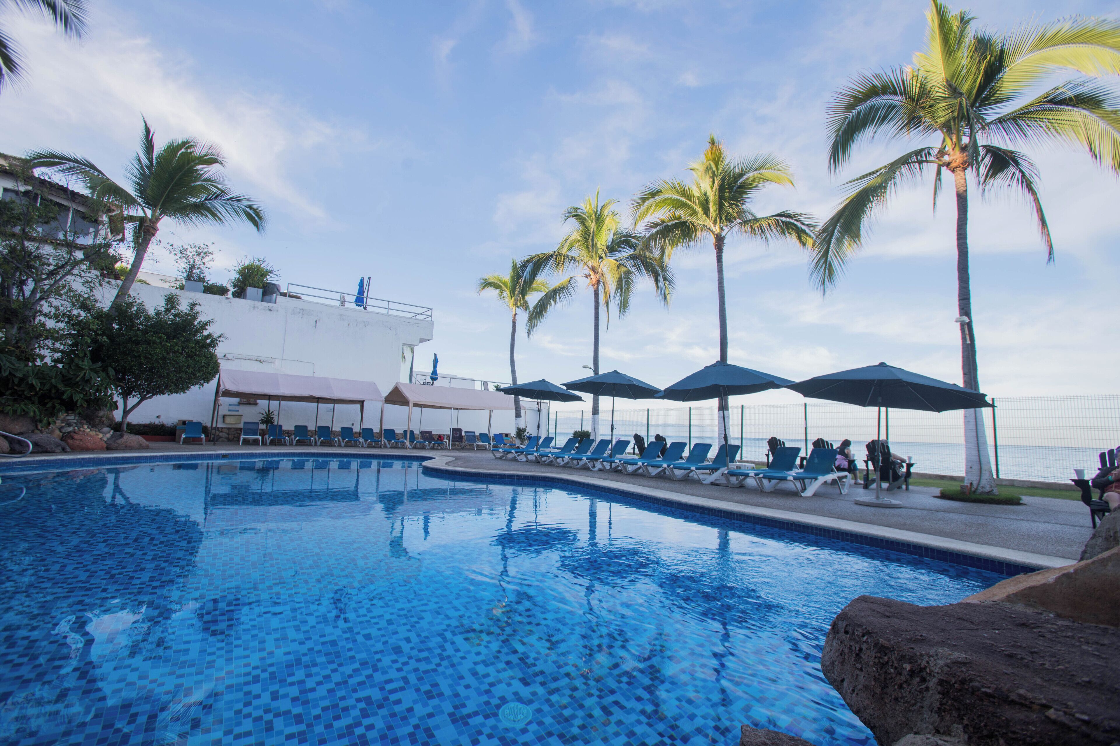 Ein Pool mit klarem, blauem Wasser, umgeben von Liegestühlen und Sonnenschirmen, mit Palmen und einem Gebäude im Hintergrund.