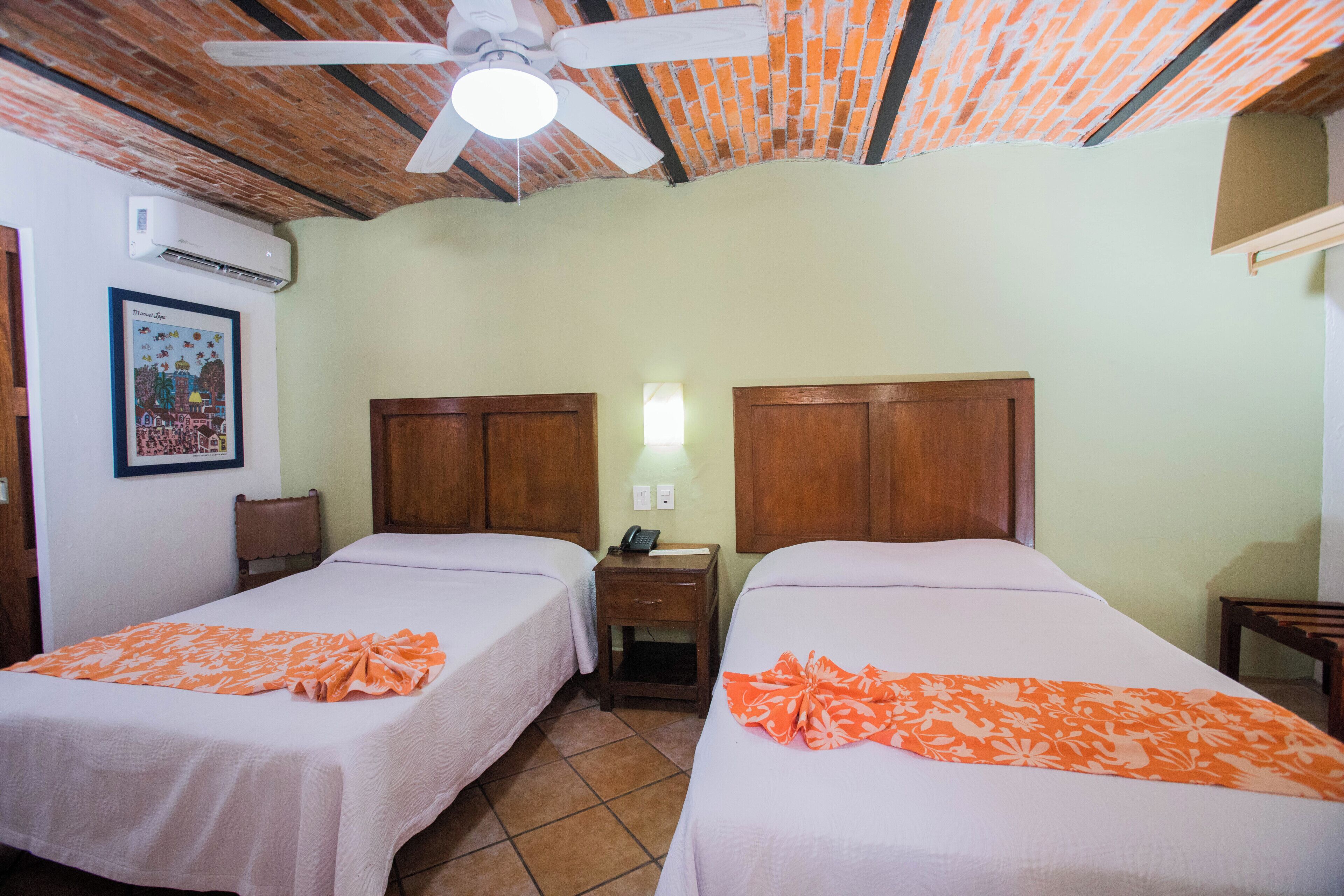 A room with two beds, a ceiling fan, an air conditioning unit, and a small bedside table with a lamp.