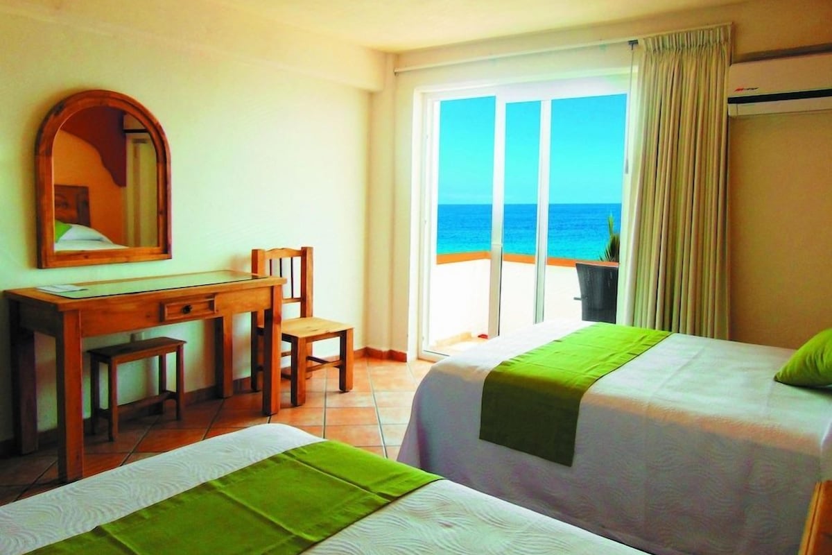A hotel room with two beds, a desk, a chair, and a large window with a view of the sea.