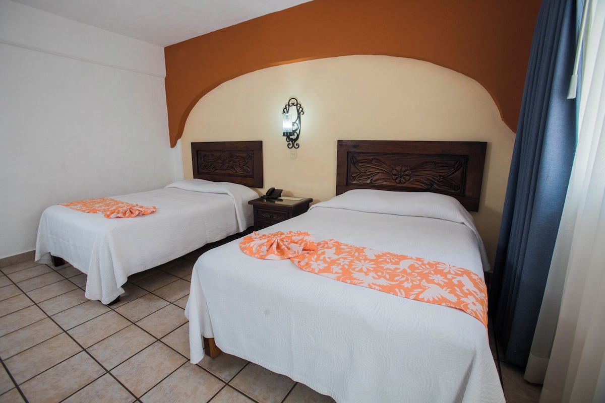 Habitación de hotel con dos camas, una mesita de noche, una lámpara de pared y una cortina.