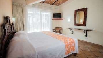 Habitación de hotel con cama, televisor montado en la pared, una silla y ventana con cortinas.