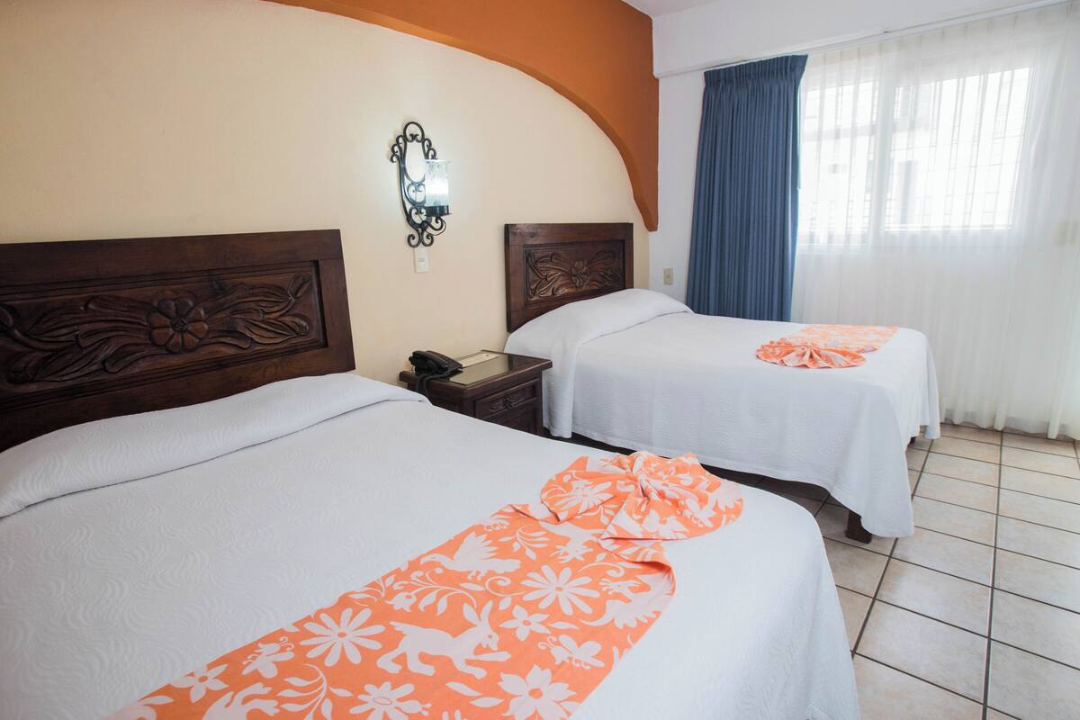 Dos camas con sábanas blancas y colchas florales naranjas, un cabecero de madera con adornos florales, una mesita de noche con un teléfono y una ventana con cortinas azules.