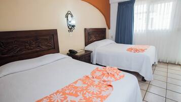 Two beds with white linens and orange floral bedspreads, a wooden headboard with floral carvings, a nightstand with a phone, and a window with blue curtains.
