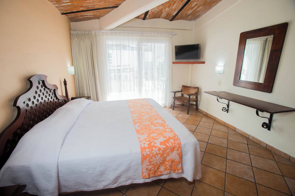 Un dormitorio con cama, una silla, un televisor, un espejo y una ventana con cortinas.