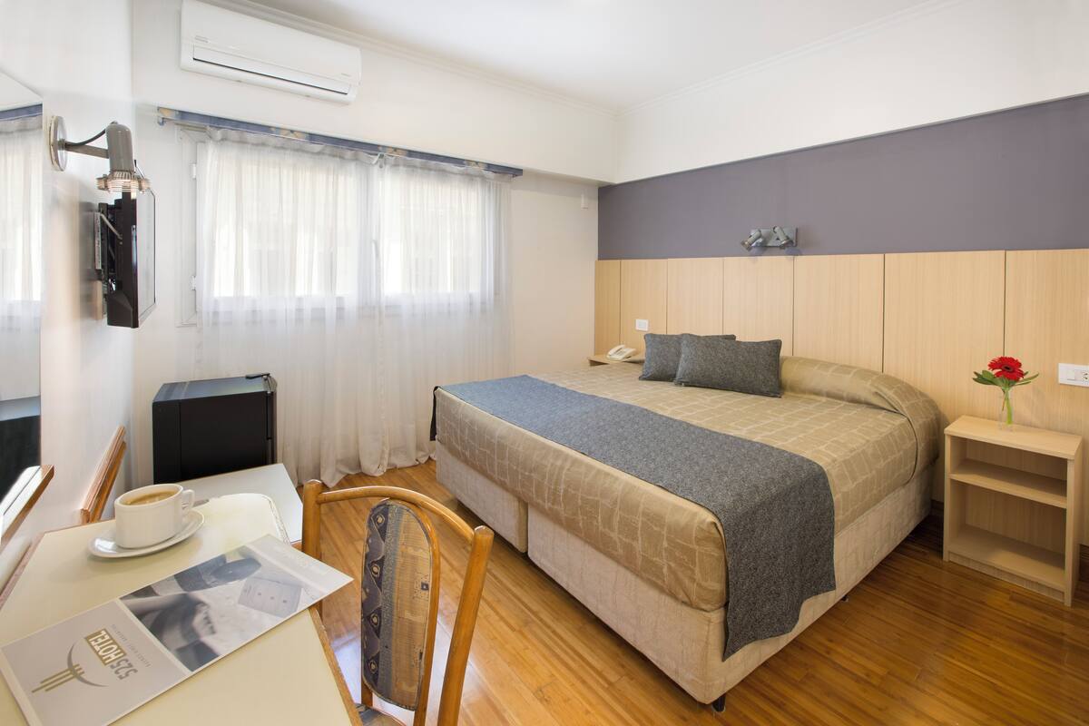 A hotel room with a bed, a desk with a chair, a coffee cup, a telephone, and a small table with a vase of flowers.