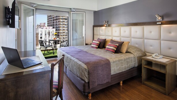 A modern hotel room with a bed, a desk with a laptop, a bedside table with a phone, and a view of the outside balcony. - Hotel Impala (Buenos Aires)