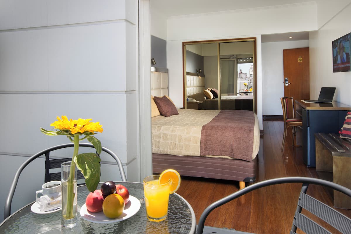 A hotel room with a bed, a desk, a chair, and a table with fruit and a drink.