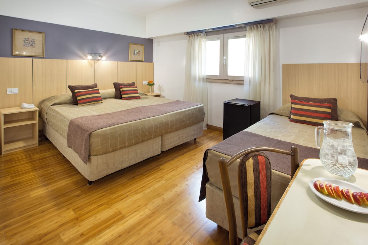 A hotel room with a large bed, bedside tables, a small table with a pitcher and fruit, and a chair.