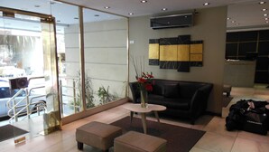 A modern interior with a glass partition, a black leather sofa, two ottomans, a small round table with a vase of flowers, and a luggage bag labeled 'FERRARO'. - Hotel Impala (Buenos Aires)