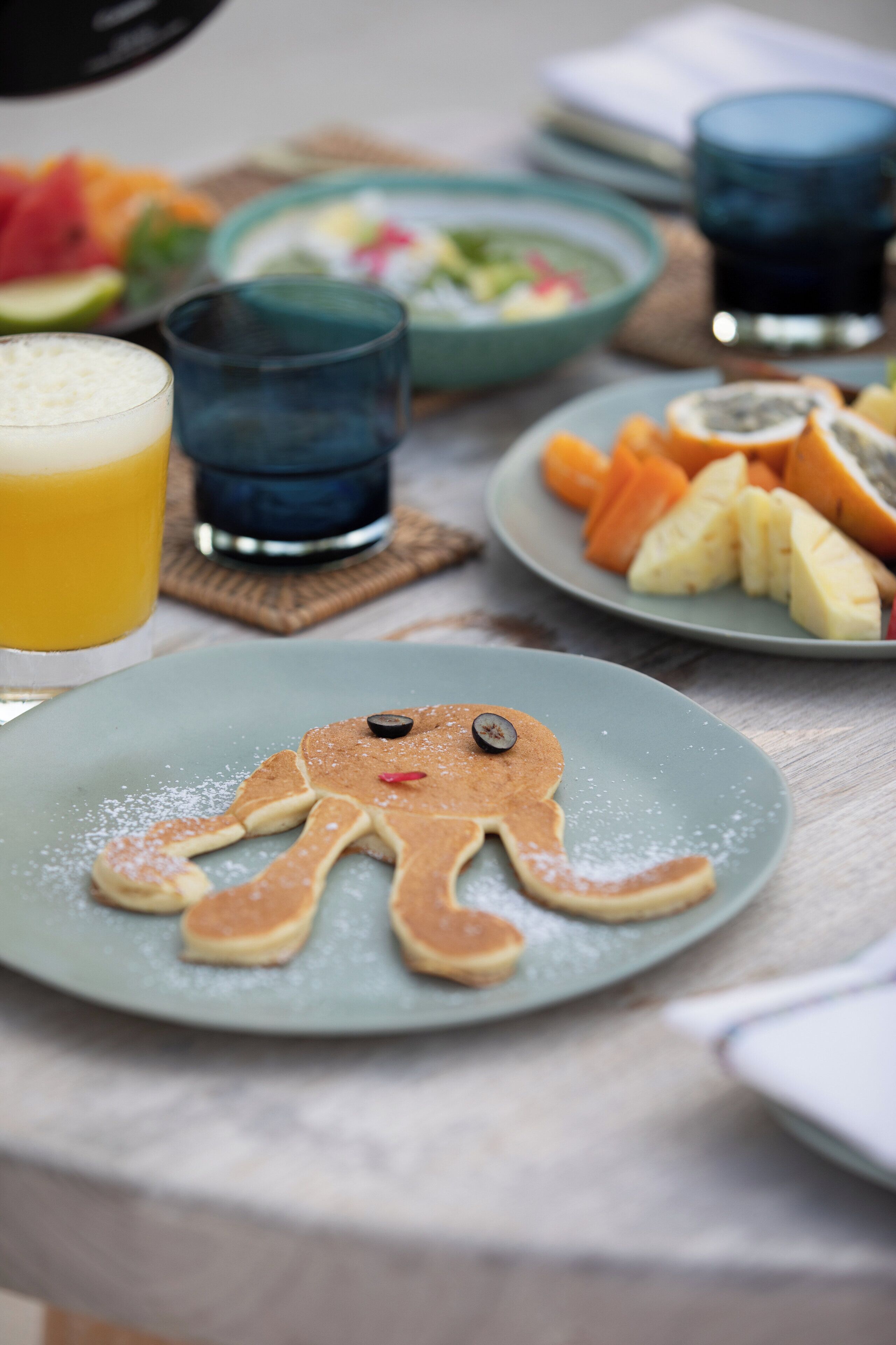A breakfast table with a plate of pancakes shaped like an octopus, a glass of orange juice, and a bowl of fruit.