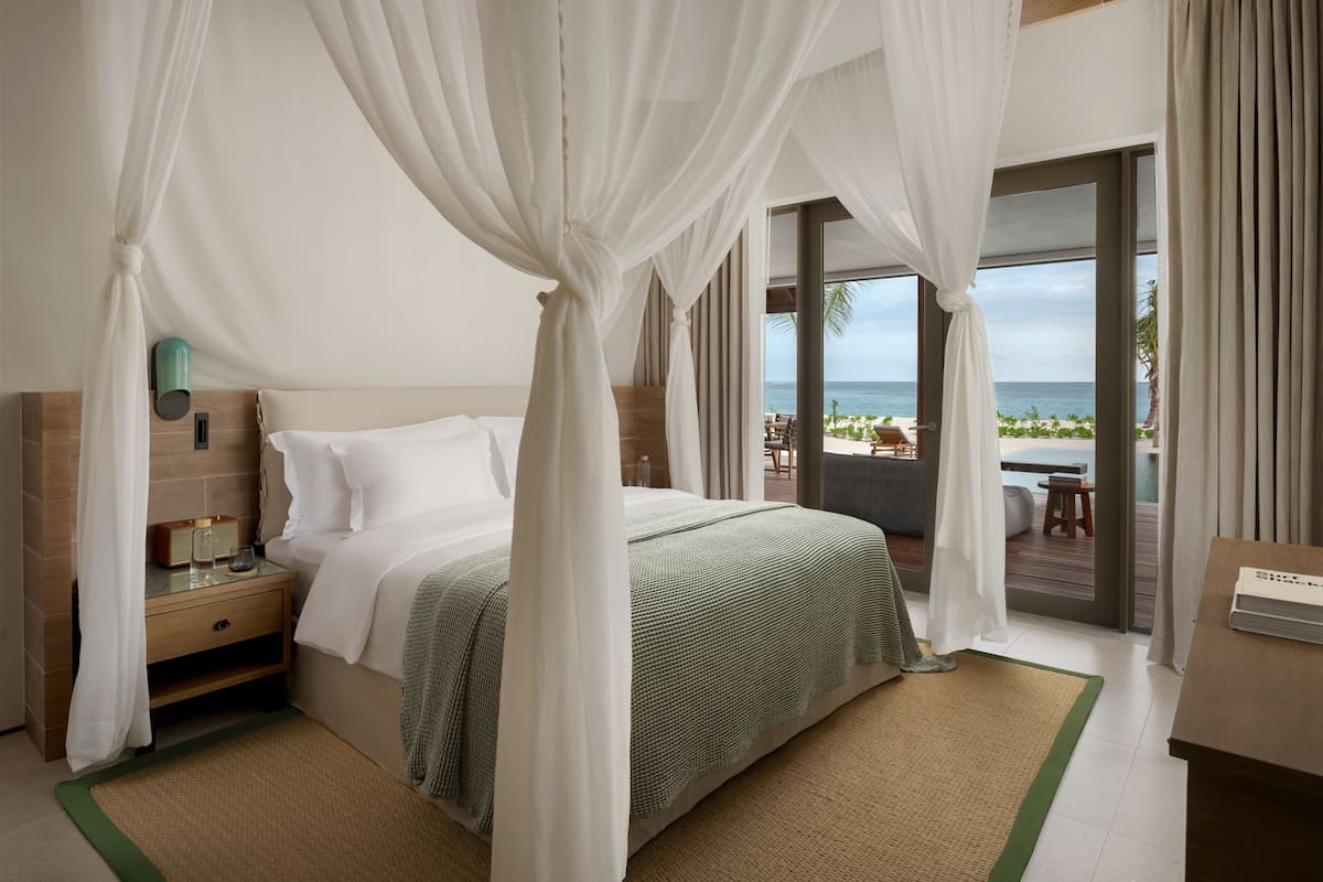Ein Schlafzimmer mit einem Bett, einem Nachttisch, einer Bettdecke in Grün und Weiß, einem gewebten Teppich und Blick auf den Strand und das Meer durch große Fenster.