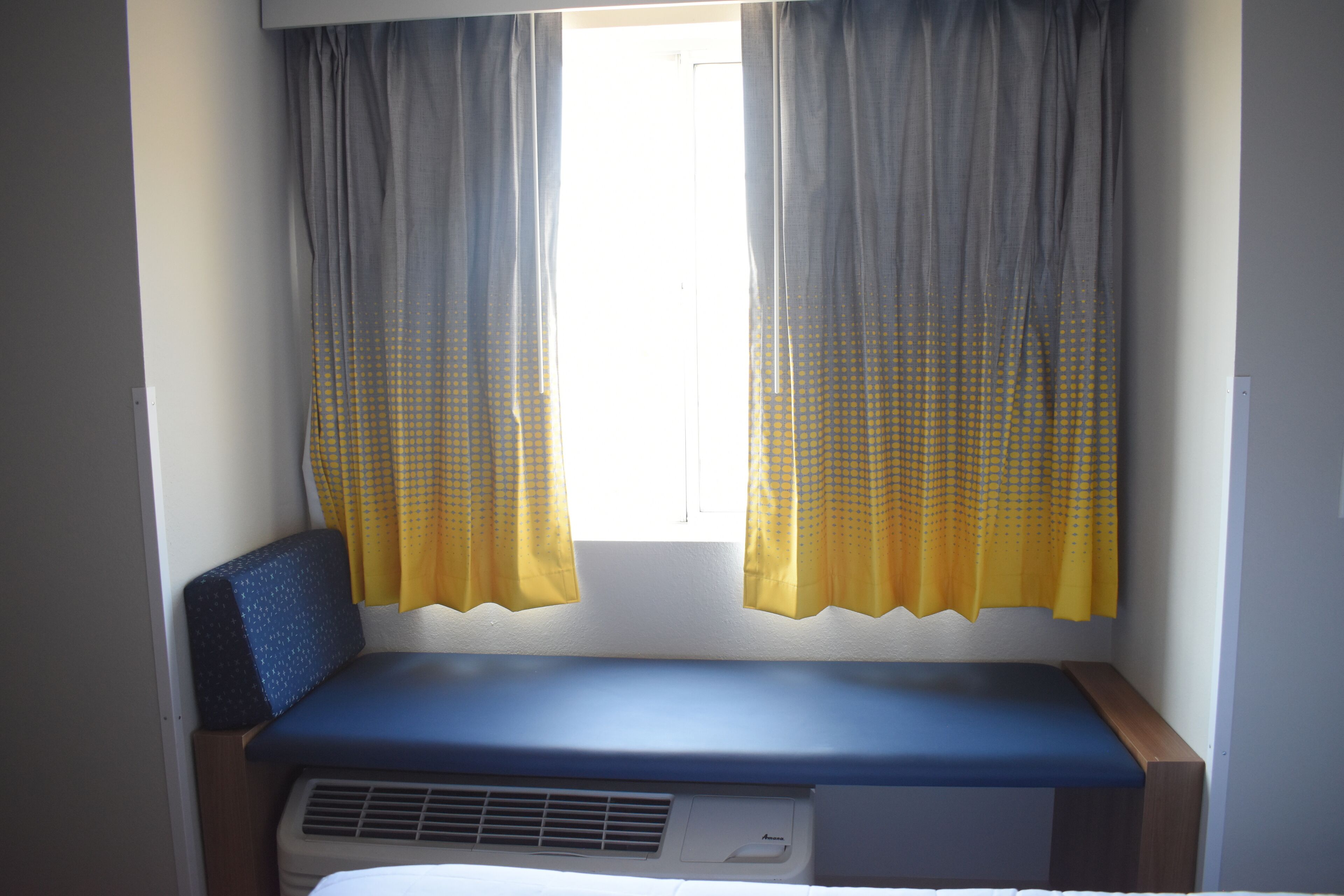 A small room with a bench, a window with curtains, and a wall-mounted air conditioning unit.