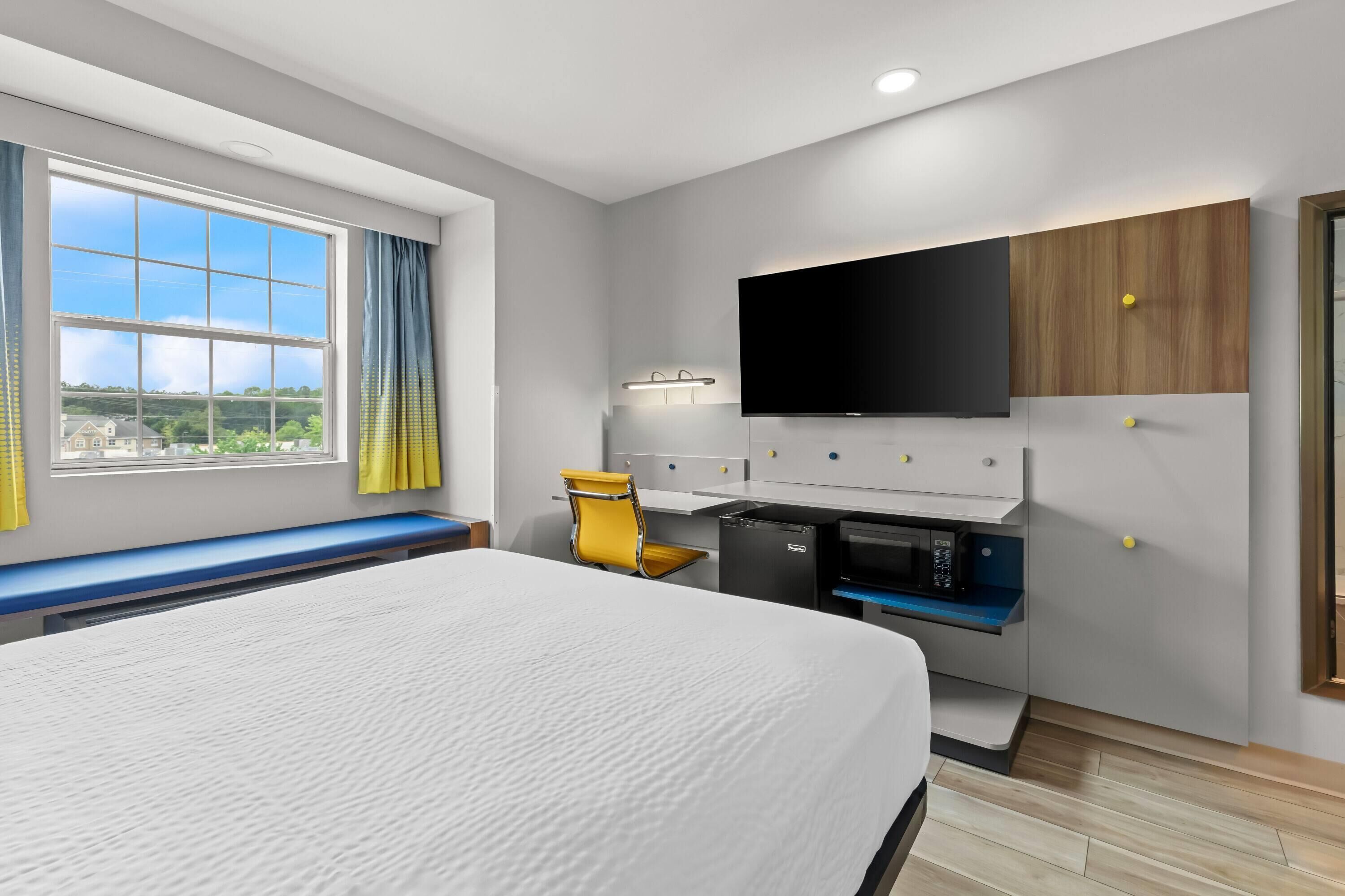 A modern hotel room with a large bed, a desk with a flat-screen TV, a microwave, and a seating area with a bench and a yellow chair.