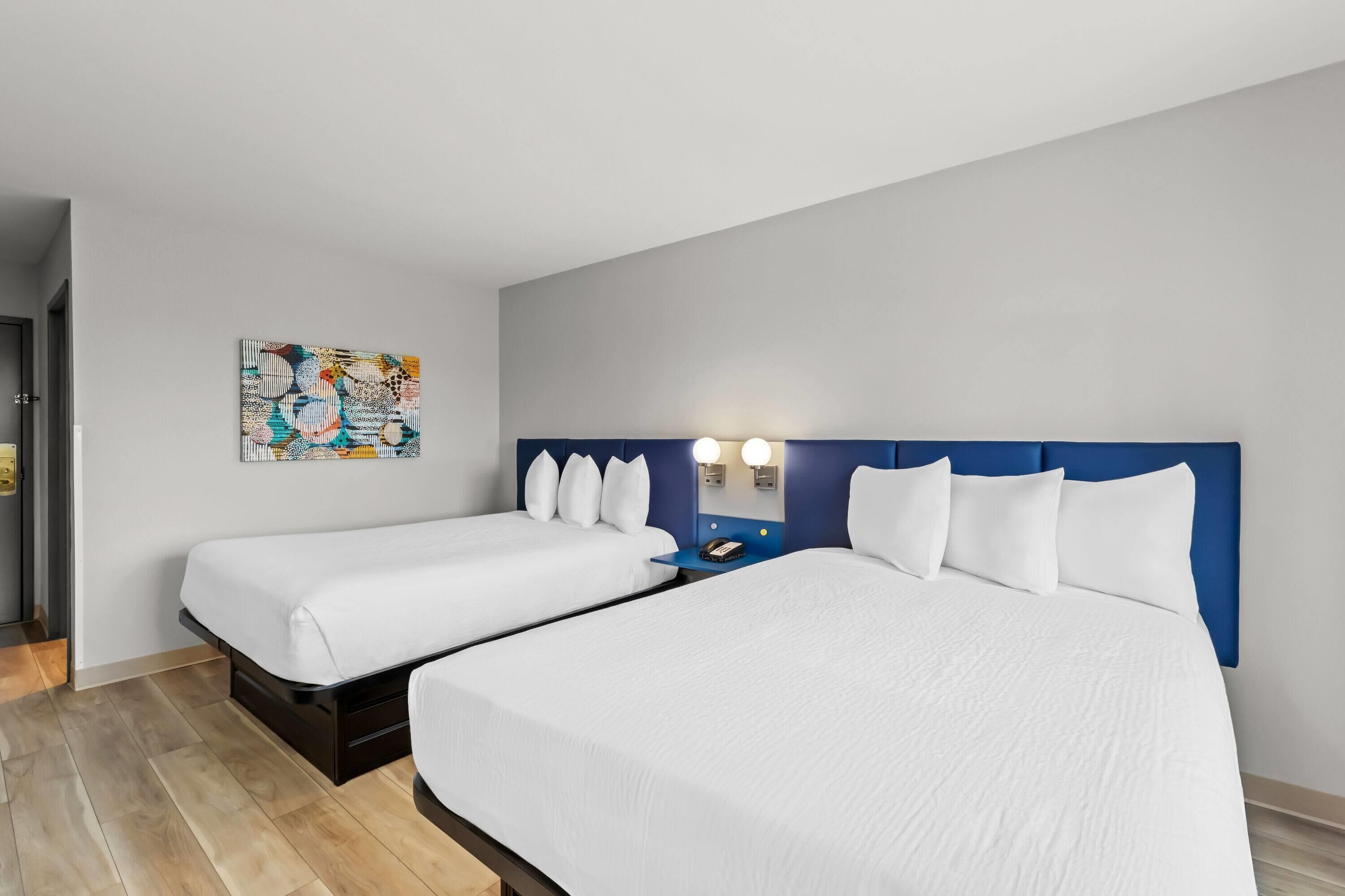 A hotel room with two beds, a blue headboard, bedside lamps, and a painting on the wall.