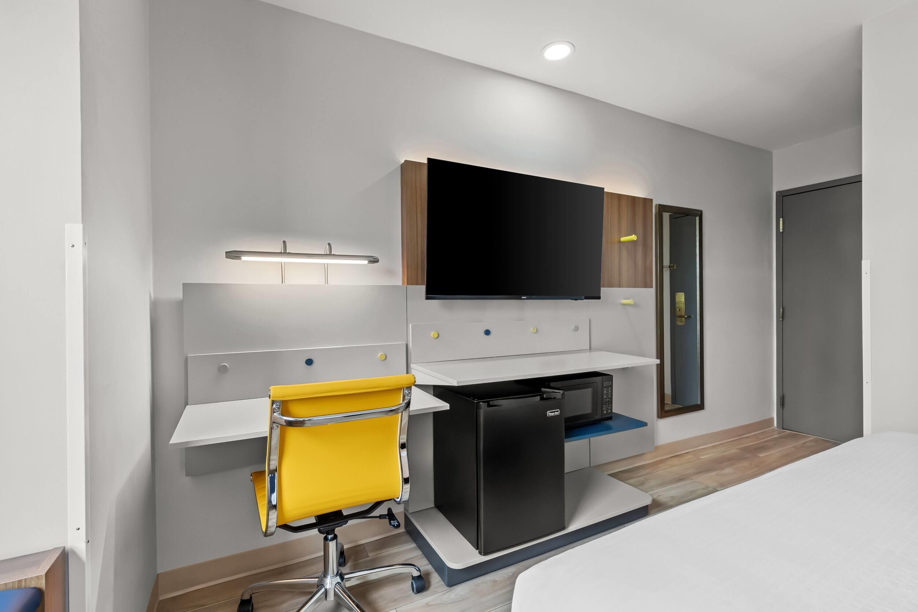 A modern hotel room with a flat-screen TV mounted above a desk, a compact refrigerator, a yellow chair, and a bed.