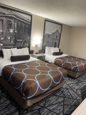 A hotel room with two beds, each with a brown and blue patterned bedspread, a nightstand with a clock, and a wall-mounted light.