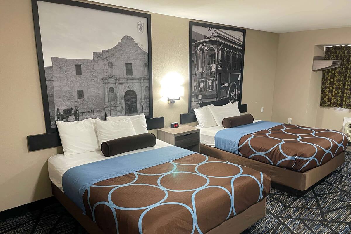 Una habitación de hotel con dos camas, cada una con un edredón estampado en tonos marrón y azul, una mesita de noche con una lámpara y un cuadro con una construcción histórica en la pared.