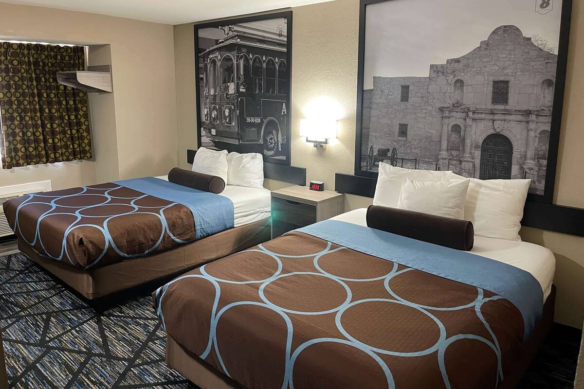 Habitación de hotel con dos camas, alfombra estampada, lámpara de pared y un cuadro con una imagen de un edificio.