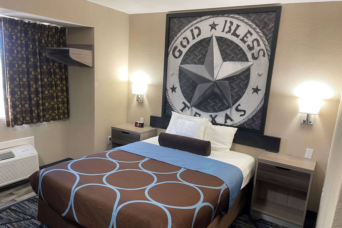 Una habitación de hotel con una cama, aire acondicionado de pared, cortina estampada, decoración de pared con una estrella y texto, y lámparas de noche.