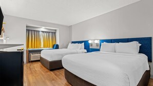 A hotel room with two beds, a desk, a window with curtains, and a blue headboard.