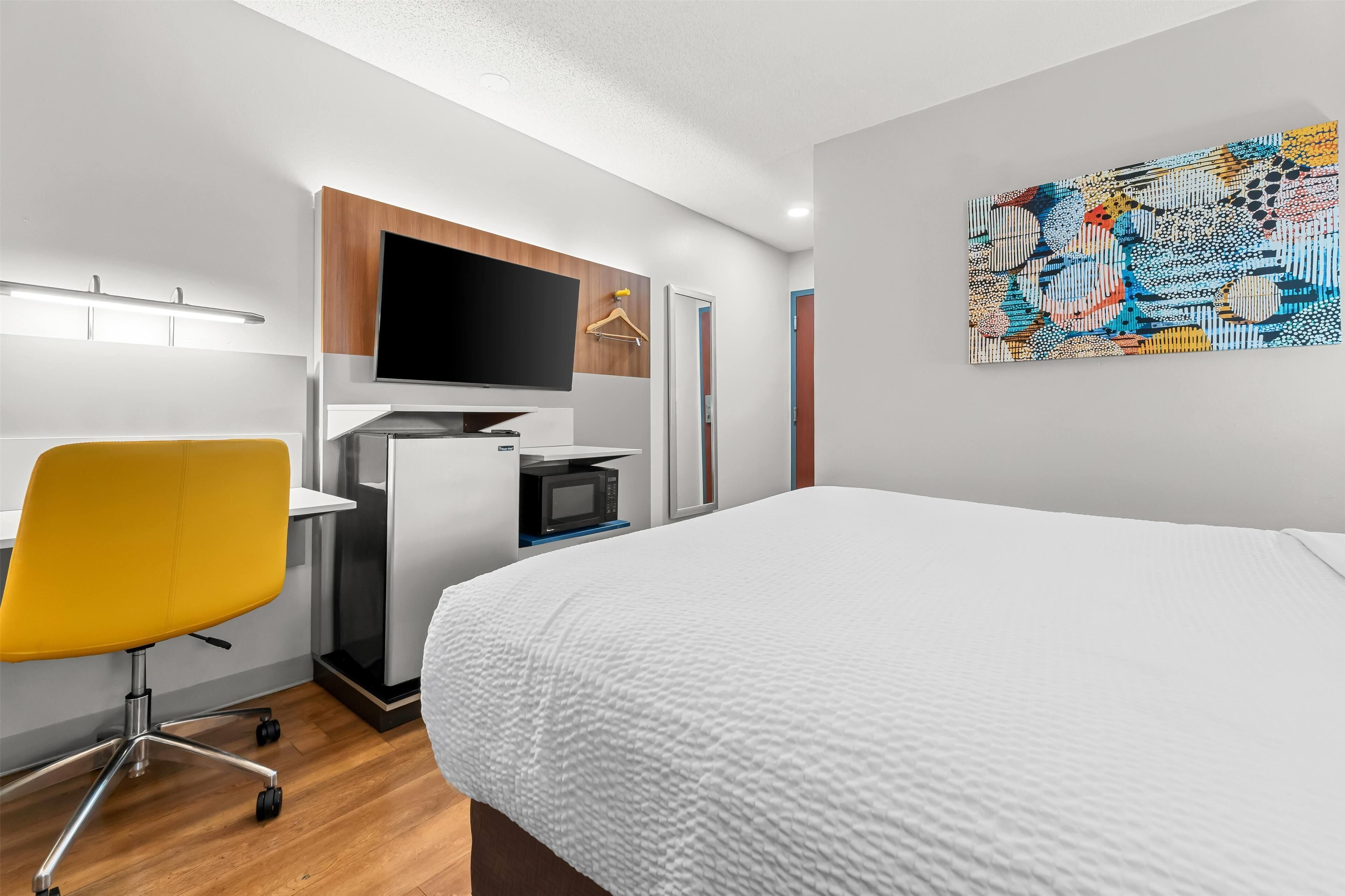 A modern hotel room with a bed, a desk with a computer, a TV, and a colorful abstract painting on the wall.