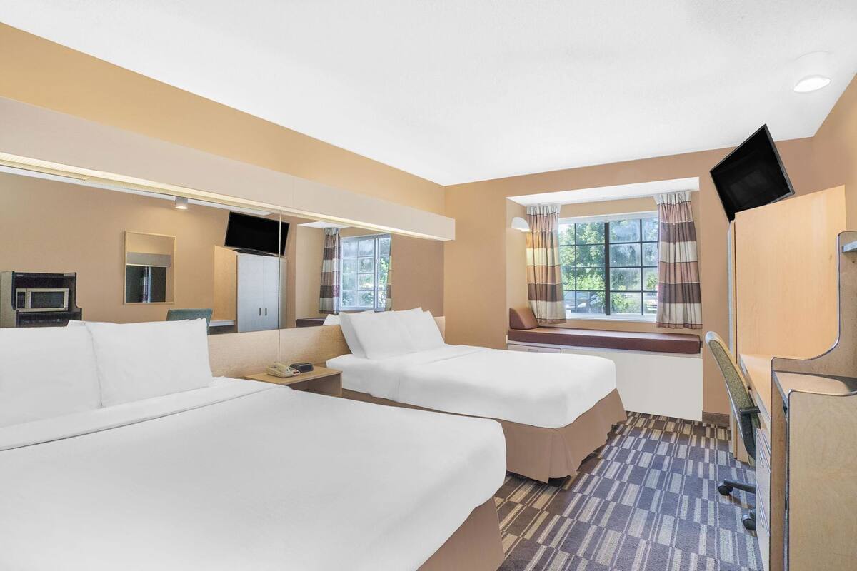 A hotel room with two beds, a TV, a window with curtains, and a flat-screen TV mounted on the wall.