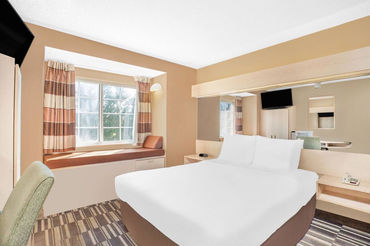 A hotel room with a large bed, a desk, a chair, a window with curtains, a flat-screen TV, and a mirror.