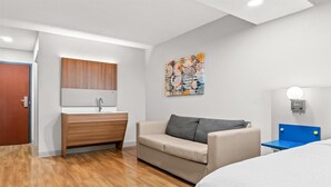 A modern hotel room with a wooden floor, a beige sofa, a small table, a bed, a wooden cabinet, a mirror, and a wall art piece.