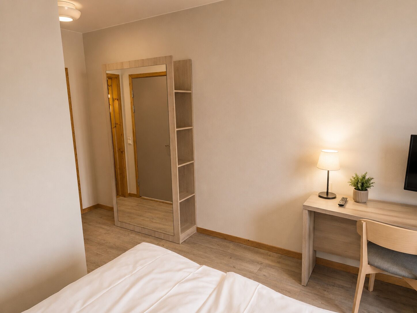 A hotel room with a bed, a desk with a lamp, a TV, and a wardrobe.