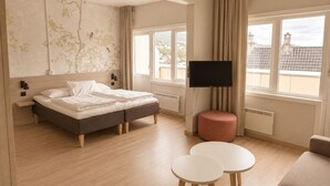 A modern hotel room with a bed, a TV, a small table, a sofa, and a view of buildings through large windows. - Haukeland Hotell (Bergen)