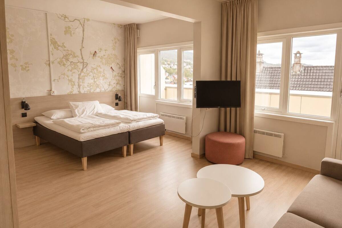 Una habitación de hotel moderna con una cama, televisor, una mesita, un sofá y vistas a edificios a través de amplias ventanas.
