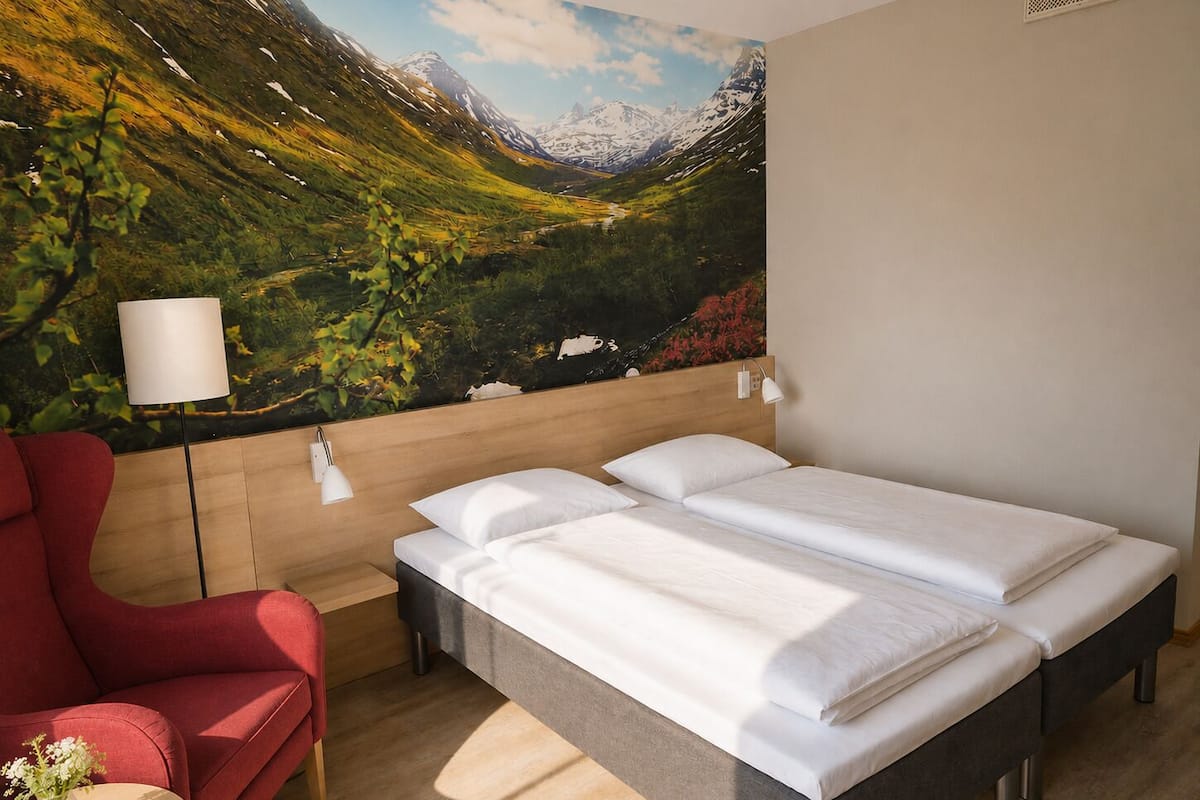 Una habitación de hotel con una cama, una silla roja, una lámpara y un mural en la pared que representa un paisaje montañoso.
