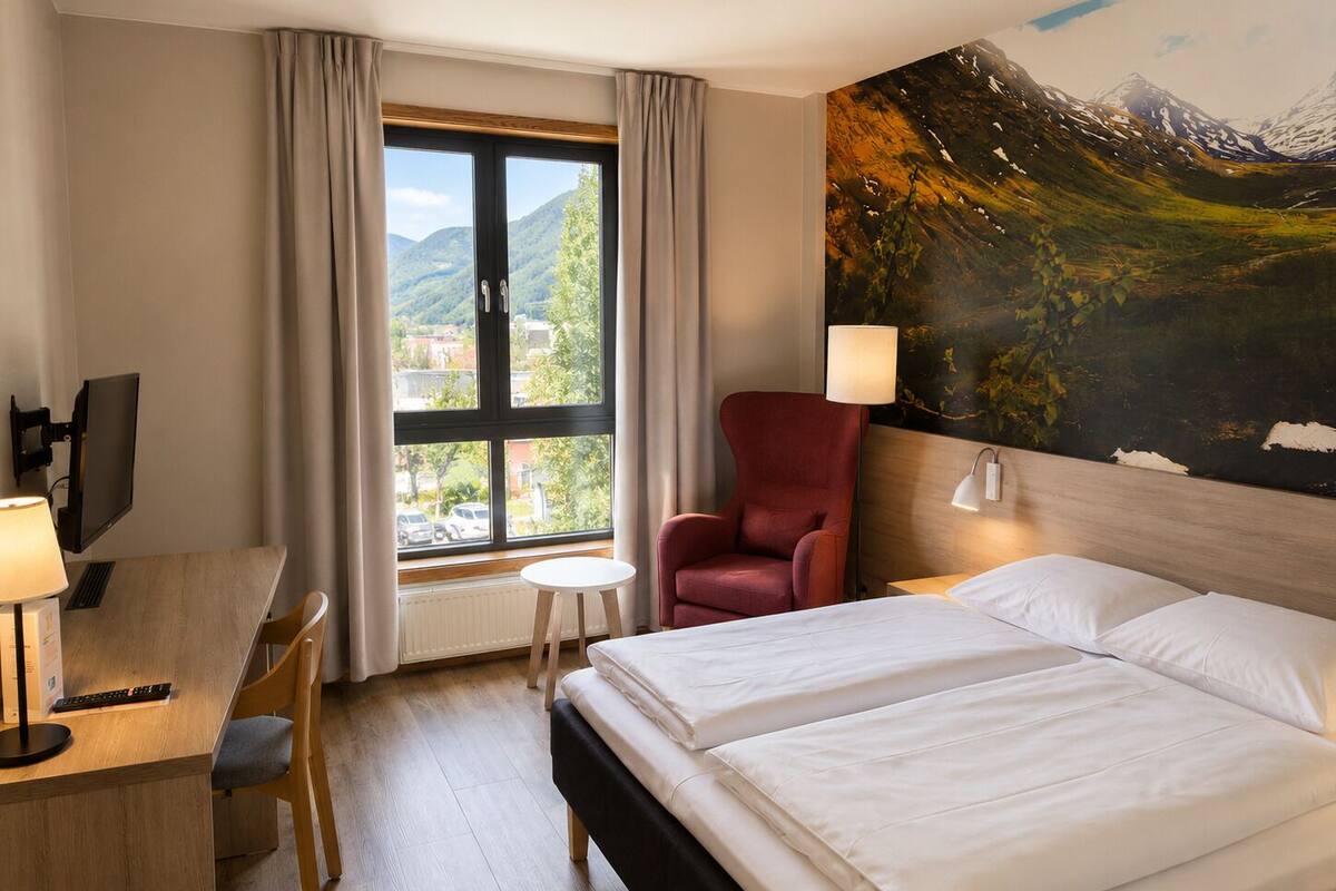 Habitación de hotel con cama, escritorio, computadora, silla y un cuadro de un paisaje montañoso.