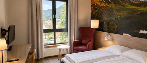 A hotel room with a bed, a desk with a computer, a chair, and a painting of a mountain landscape.