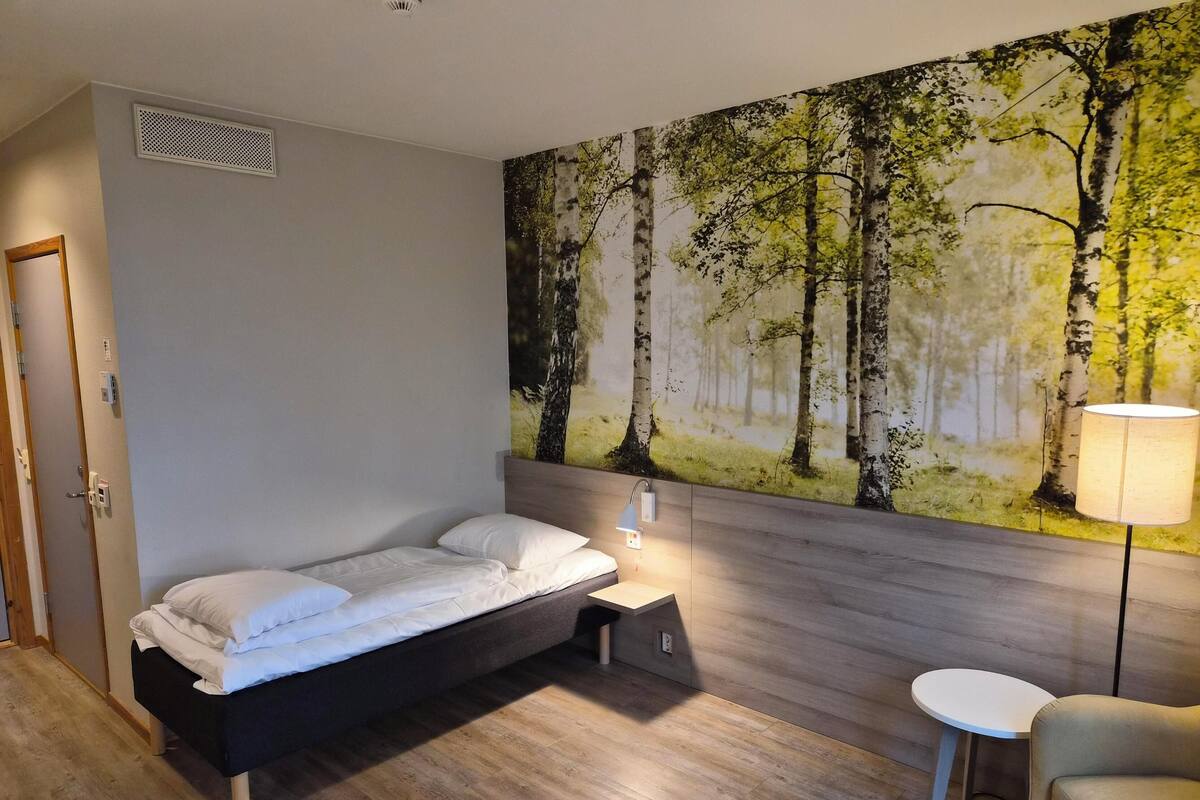 Un dormitorio con cama, mesita de noche, lámpara y un cuadro de un bosque.