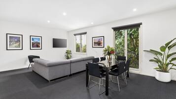 A modern room with a grey sofa, a dining table with chairs, a flat-screen TV mounted on the wall, and large windows with a view of greenery.