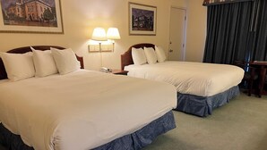 A hotel room with two large beds, a nightstand with a lamp, and a door with a curtain. - Hardman House Inn & Suites (Carson City)