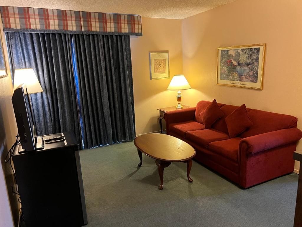 A hotel room with a red sofa, a wooden coffee table, a desk with a lamp, and a painting on the wall.