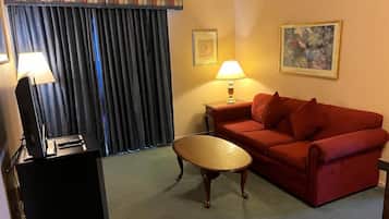 A hotel room with a red sofa, a wooden coffee table, a desk with a lamp, and a painting on the wall.
