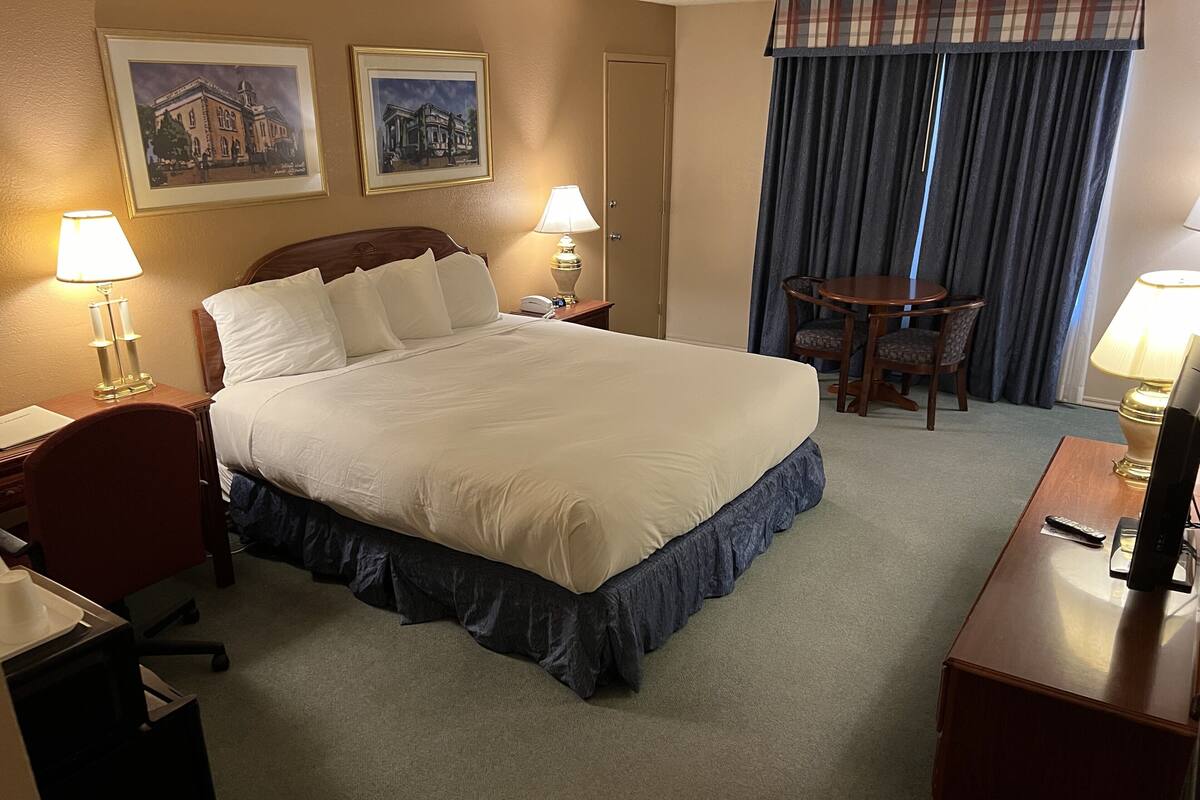 A hotel room with a large bed, two bedside lamps, a desk with a chair, a small table with a chair, and two framed pictures on the wall.