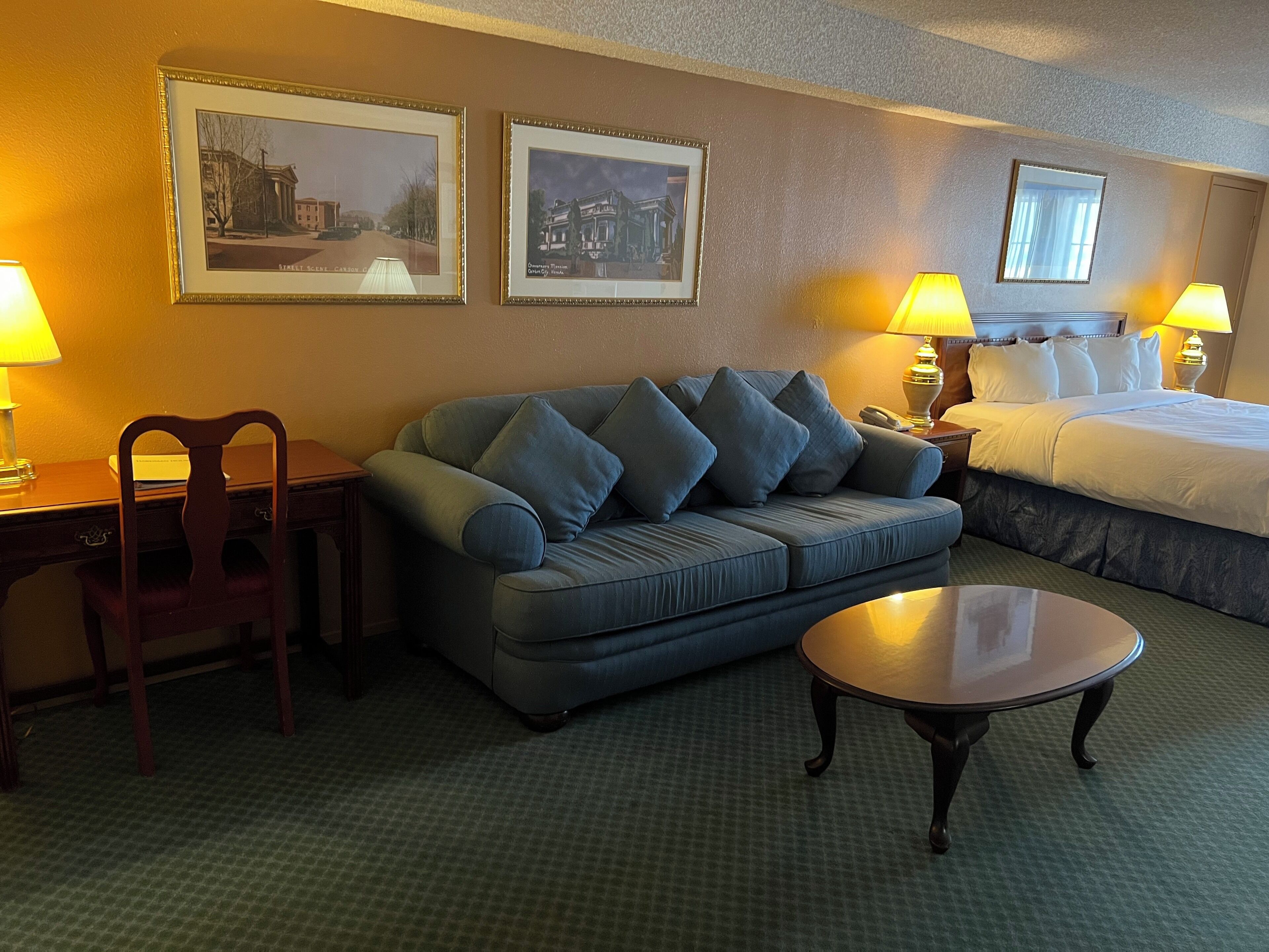 Habitación de hotel con sofá, una silla, una mesita, cama y dos lámparas. Se observan dos cuadros en la pared.