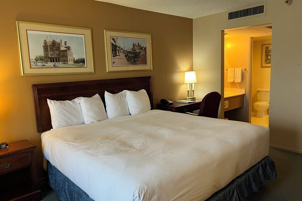 A hotel room with a large bed, a desk with a lamp, and two framed pictures on the wall.