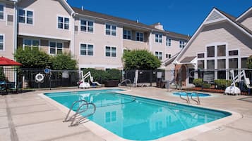 A swimming pool with a surrounding fence, a red umbrella, and a building with multiple windows and a sloped roof.