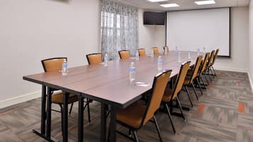 Un salón de conferencias con una larga mesa, sillas, botellas de agua, una pantalla de proyector y un equipo de aire acondicionado.