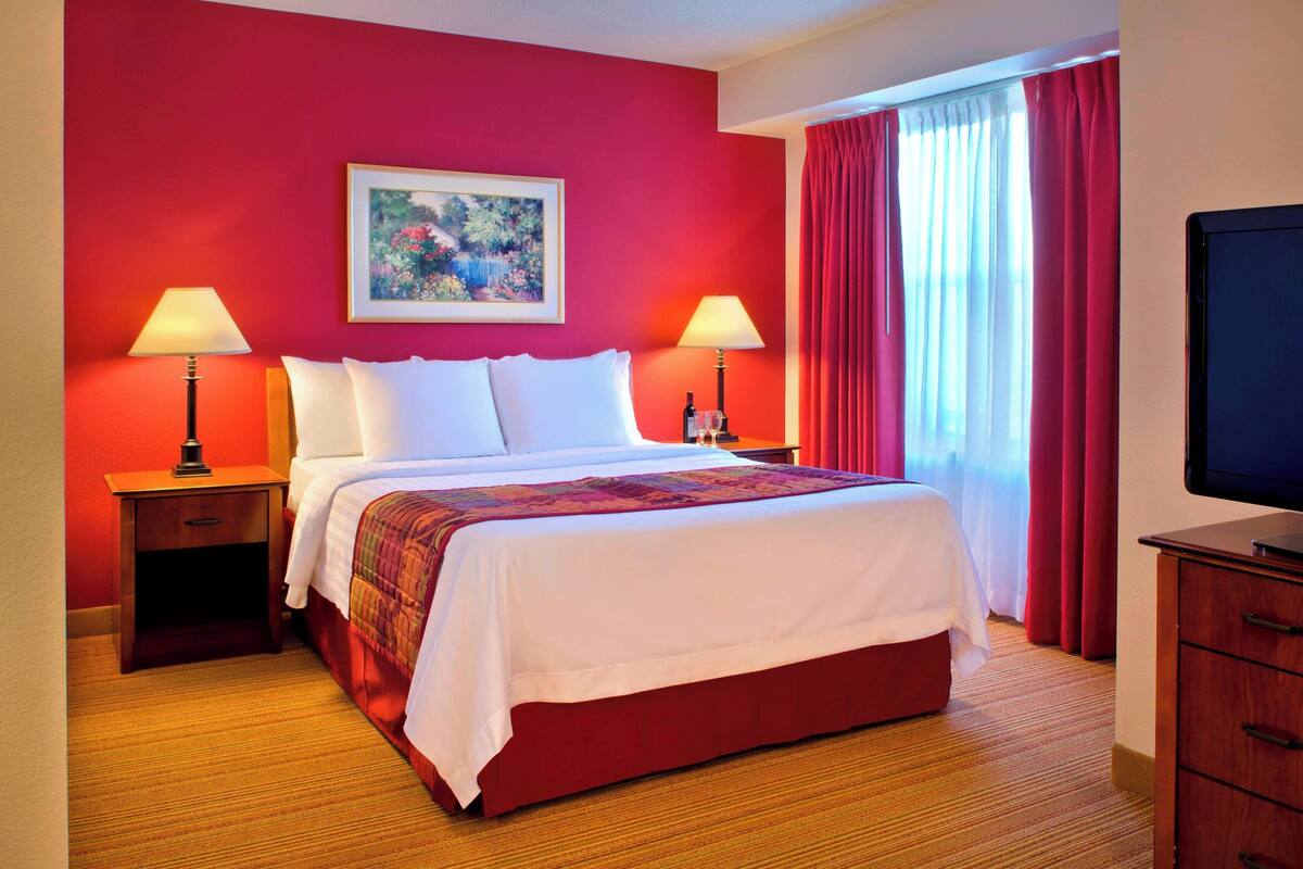 A hotel room with a large bed, two bedside tables with lamps, a television, and a window with red curtains.