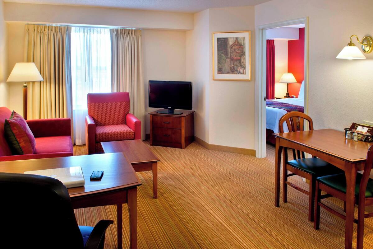 A hotel room with a red sofa, a wooden dining table, a television, and a bed with a red wall and a lamp.