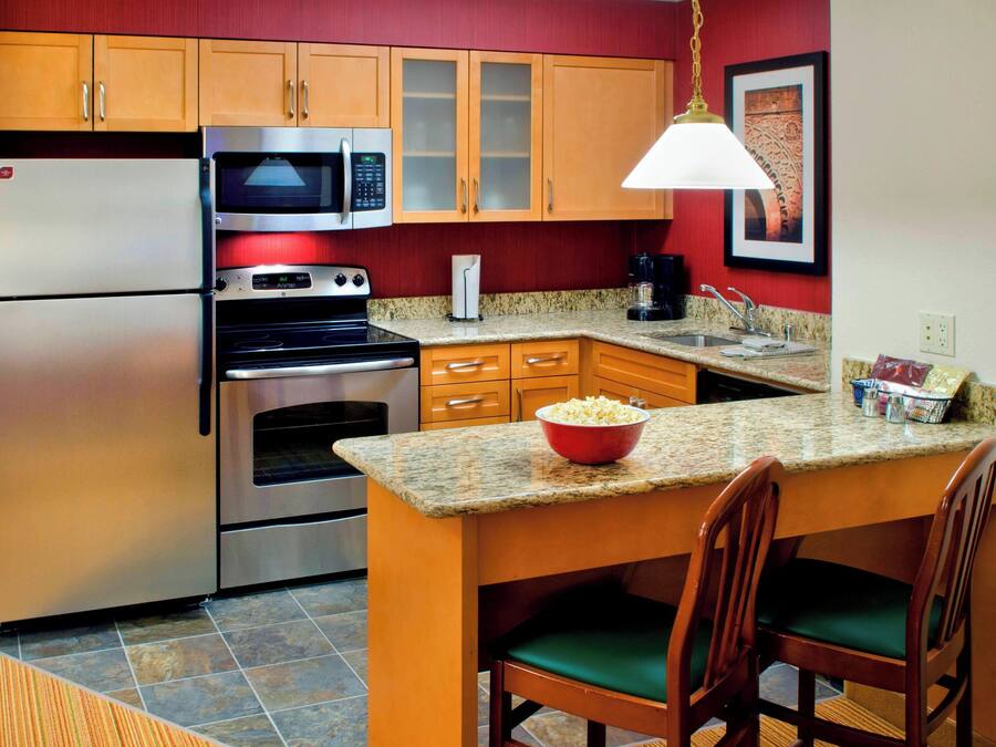 A kitchen with a refrigerator, microwave, oven, granite countertop, wooden cabinets, and a dining area with chairs.